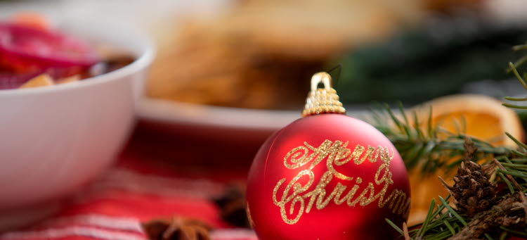 En röd julkula med texten Merry Christmas i guld stående på ett julbord.