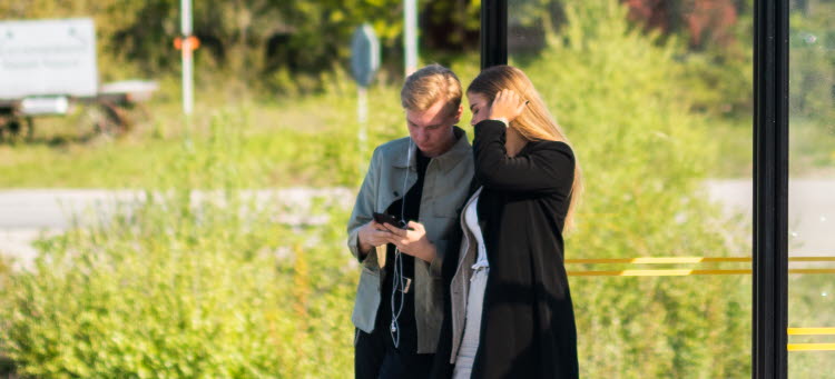 Två ungdomar står vid en hållplats och kollar på en mobiltelefon
